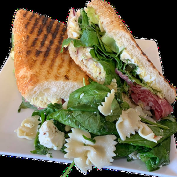 Grilled panini sandwich with greens and pasta salad on a white plate.