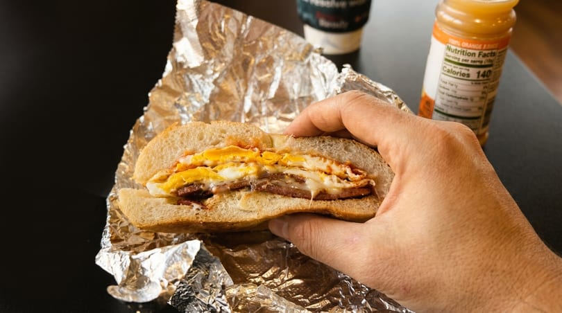 A hand holds a breakfast sandwich with egg, cheese, and meat, wrapped in foil, with orange juice in the background.