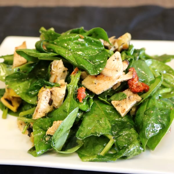 Spinach salad with grilled chicken, sun-dried tomatoes, and yellow squash on a white plate.